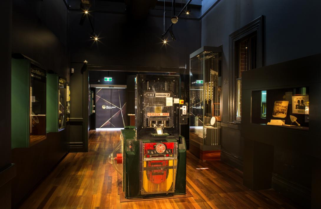 A vintage coin press 'HME coinmaster 2', painted green and red, sits in the centre of a museum gallery with display cases along the walls on either side.