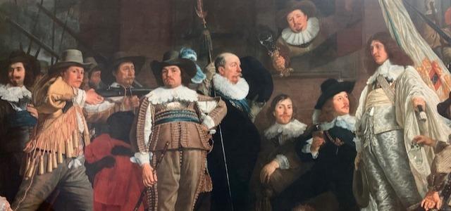 A painting showing a group of seventeenth century Dutch men. They are finely dressed in hats, with lace collars and sashes. Some hold weapons, some look at the viewer, and others look off to the distance.