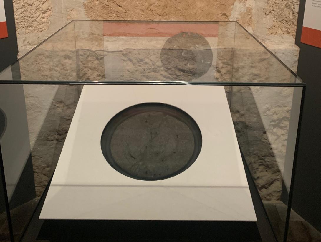 A flattened pewter plate with text inscribed on it. It sits in a square white surround within a glass display case.
