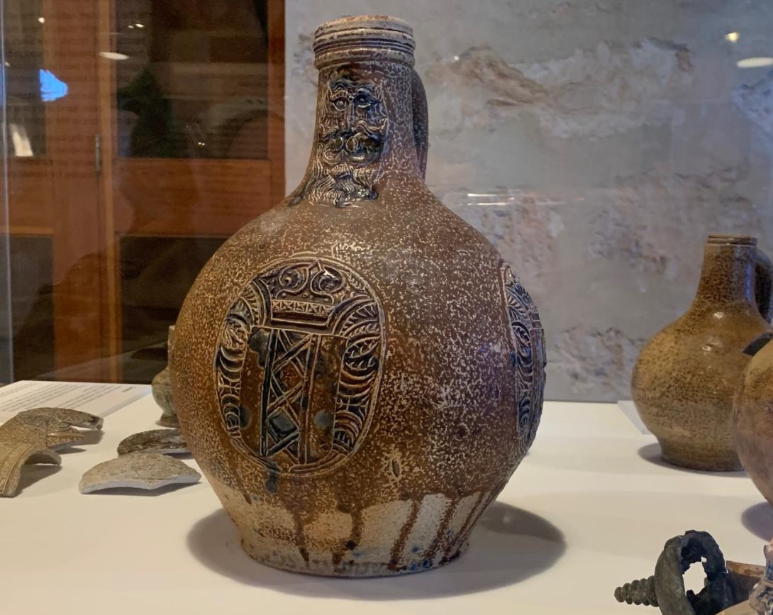 Stoneware jug featuring a bearded face design on the neck of the vessel, and a medallion with blue detail on the belly of the jug.