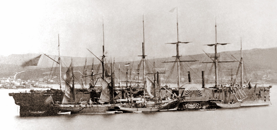 Black and white photo of large ship with many masts. Four smaller ships sit alongside the larger ship. There are buildings and hills pictured in the distance