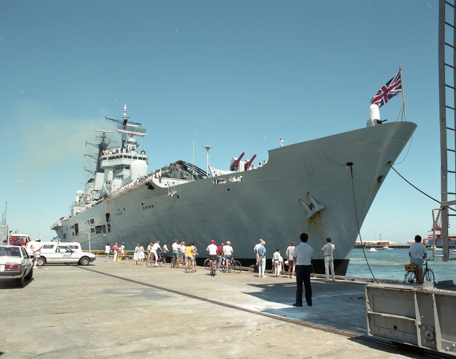 Colour photograph of a large naval ship docked at a wharf on a clear, sunny day. The ship flies a maritime flag at its bow and towers above a crowd of people gathered on the pier. Visitors stand, walk, and ride bicycles along the waterfront while looking up at the vessel. Cars and service vehicles are parked nearby, and the calm harbour water reflects the bright blue sky.