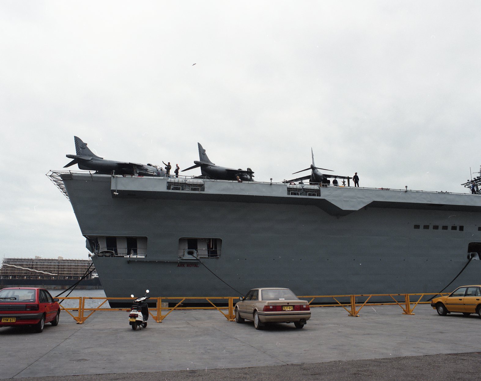 Colour photograph of the bow section of an aircraft carrier moored at a wharf. Several jet aircraft are parked along the forward edge of the flight deck, with crew members walking and working nearby. Below the overhang of the deck, the ship’s grey hull rises high above the waterline. Cars and a scooter are parked on the wharf in the foreground, and the sky is overcast.