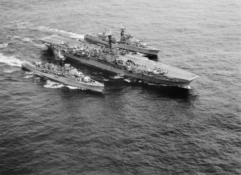 A black‑and‑white aerial photograph showing three naval vessels traveling side by side across open water. In the center is a large aircraft carrier with aircraft and equipment visible on its deck. A smaller warship sails tightly along each side of the carrier, creating parallel wakes as the three ships move forward in formation.