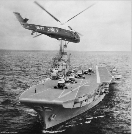 Black‑and‑white photograph of an aircraft carrier at sea with several helicopters parked on its flight deck. Another helicopter hovers just above the ship, aligned over the deck with its rotors spinning. Crew members stand near the parked aircraft, and the ocean stretches into the distance under a clear horizon.