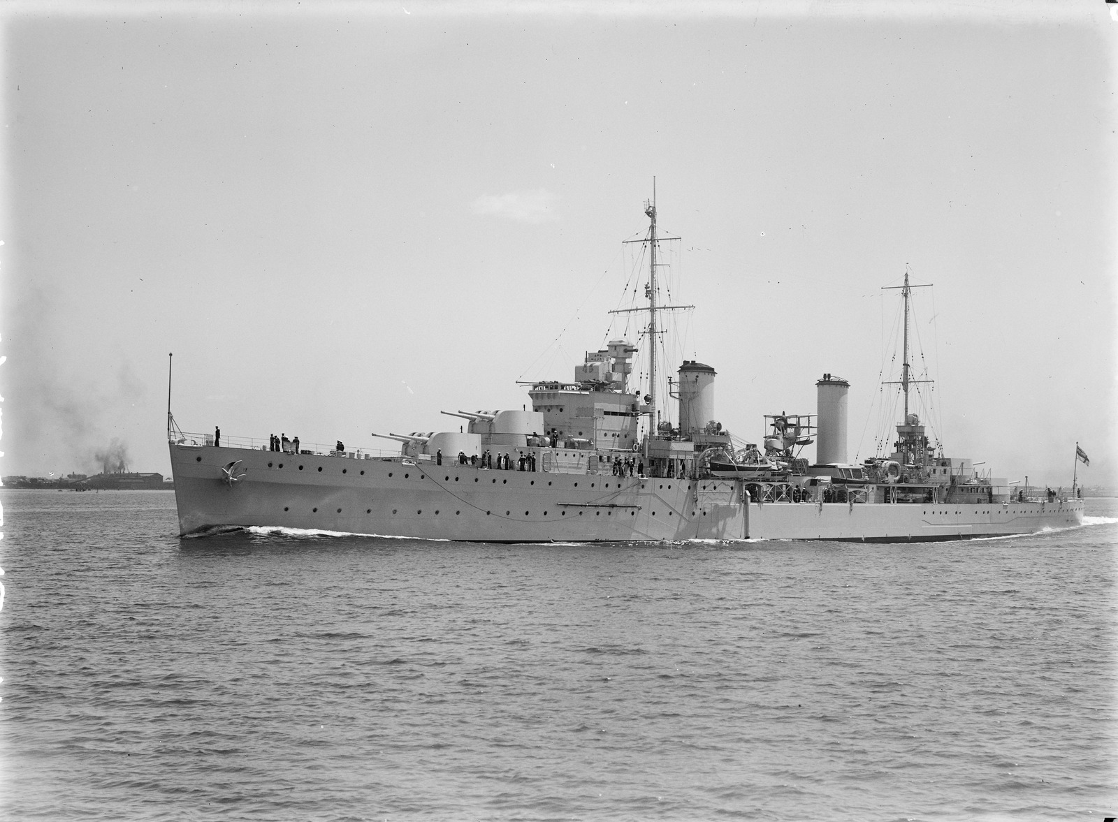 Black‑and‑white photograph of a large naval warship traveling across calm water, viewed from the side. The ship has two tall masts, multiple gun turrets, and a long, sleek hull. Crew members are visible standing along the decks. Smoke can be seen rising from another vessel far in the background on the left, while the foreground ship moves steadily forward under a clear sky.