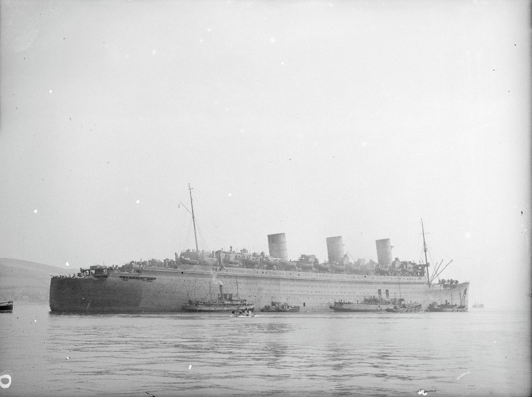 Black‑and‑white photograph of a large ocean liner converted for use as a troopship, anchored in a calm bay. The ship has a long hull and three tall funnels, with lifeboats and fittings mounted along the upper decks. Several small craft and tugboats cluster around the vessel near the waterline. The distant shoreline is faintly visible through light haze under an overcast sky.