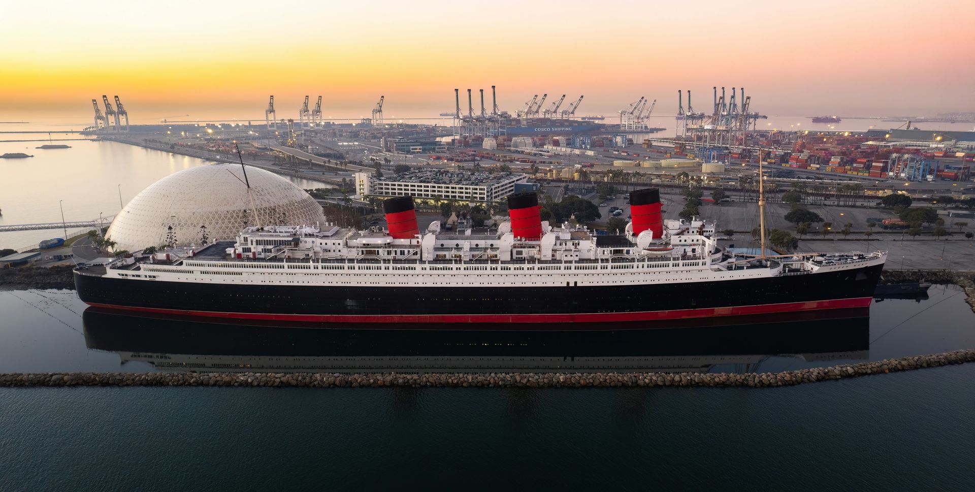 A wide, high‑angle colour photograph of the historic ocean liner Queen Mary docked in calm harbour waters at sunset. The ship’s three red‑and‑black funnels stand out along its long white superstructure and dark hull. A curved breakwater runs parallel to the vessel, and behind it stretches a busy port filled with cranes, shipping containers, and industrial buildings. The sky glows with soft orange and pink tones as daylight fades over the harbour.