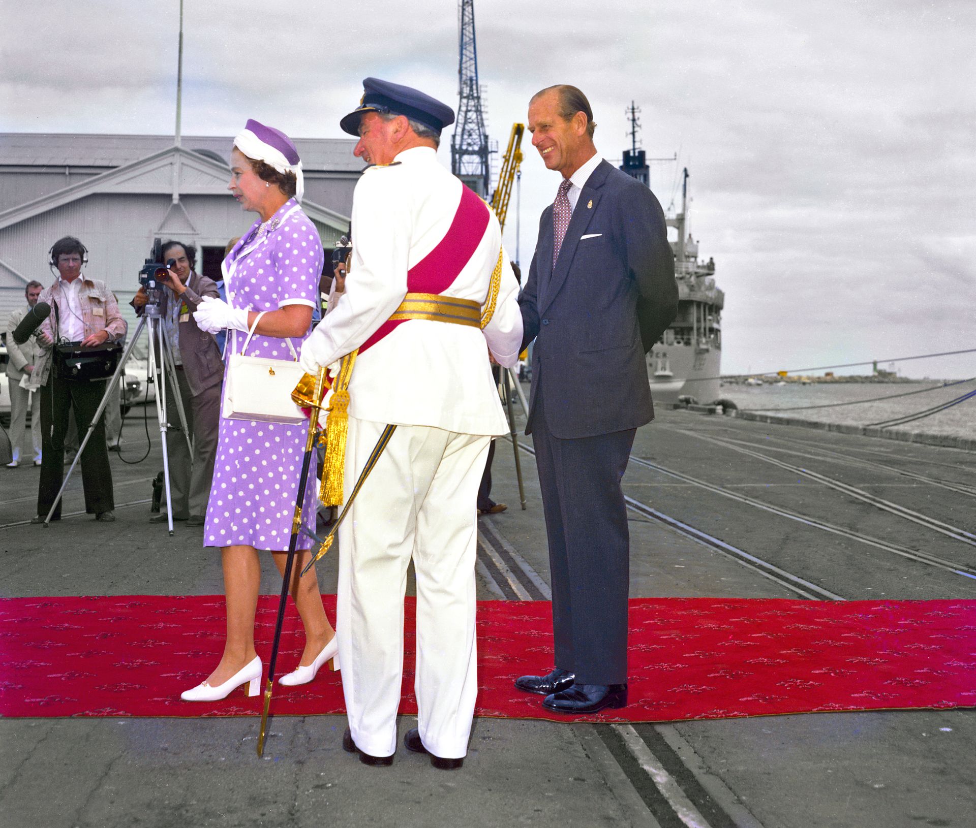 Colour photograph of three individuals standing on a red ceremonial carpet laid out on a dockside. One person wears a purple patterned dress with matching hat, gloves, and handbag. Another person beside them is dressed in a dark formal suit with hands clasped behind their back. A third person, wearing a white dress uniform with a blue cap and ceremonial sash, stands facing them while holding an ornate staff. Photographers and onlookers stand in the background near buildings and harbour structures, and a lar