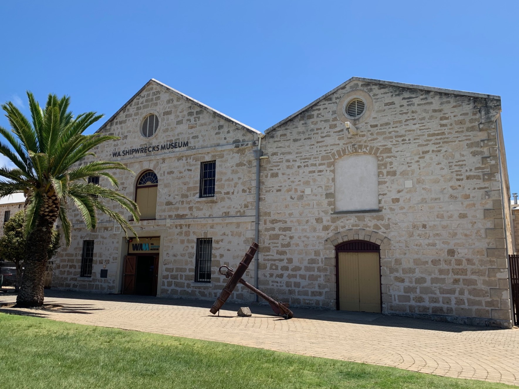 A two-story limestone building, with a paved entry way. A large anchor sits on the pavement. In front of the building is a large palm tree, and a green grass lawn.