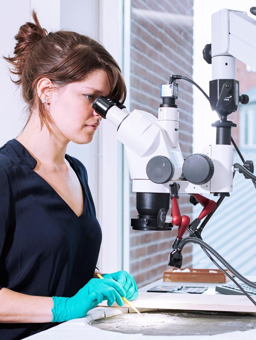 Woman wearing rubber gloves and holding a small brush, standing looking through a microscope at a metal disc.
