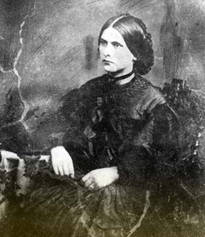 Old black and white portrait photo of a woman seated, not smiling with her hands in her lap, and hair wrapped around with a bun at the back.