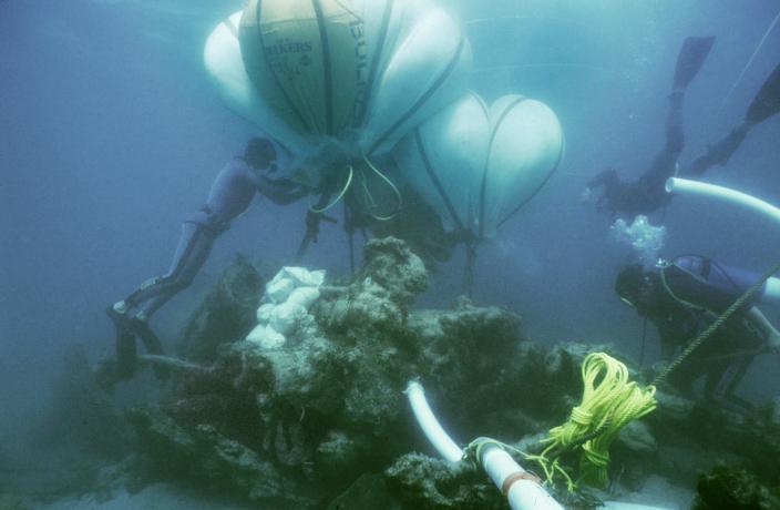 Underwater image of a inflated bags attached to a piece of wreckage on the seabed, with two divers manouvering the airbags and ropes.