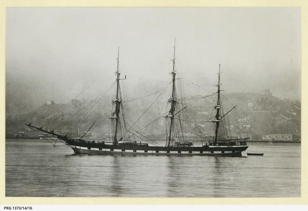 A three-masted ship at anchor on still water. In the background a hilly town can be seen behind a shroud of mist.