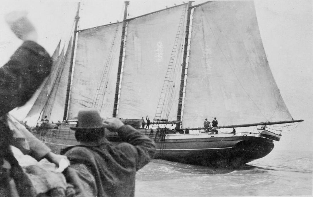 Men on shore wave at the ship loaded with timber, its sails now up.