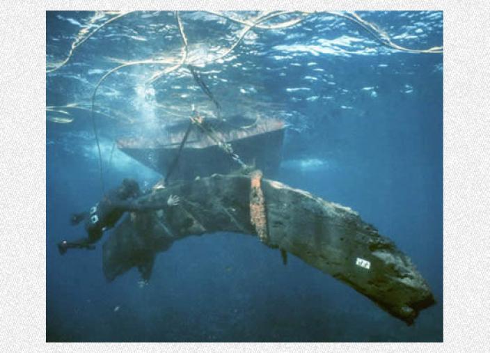 A very large, curved section of Batavia's timbers underwater, being raised up to the water's surface. A Museum diver guides it into position and the Museum's boat's underside is visible behind.