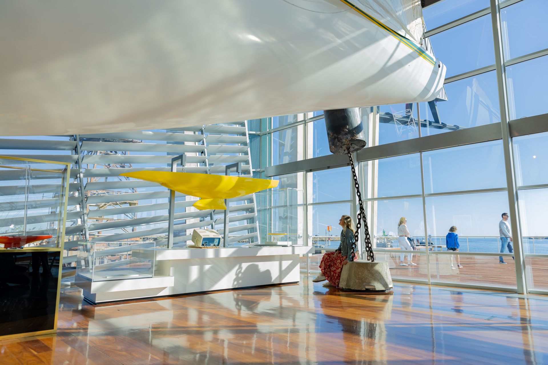 A large brightly lit gallery overlooking the water. The yacht Australia II is on one side and Jon Sanders' yacht is on the other. A stair leading to a walkway is in between.
