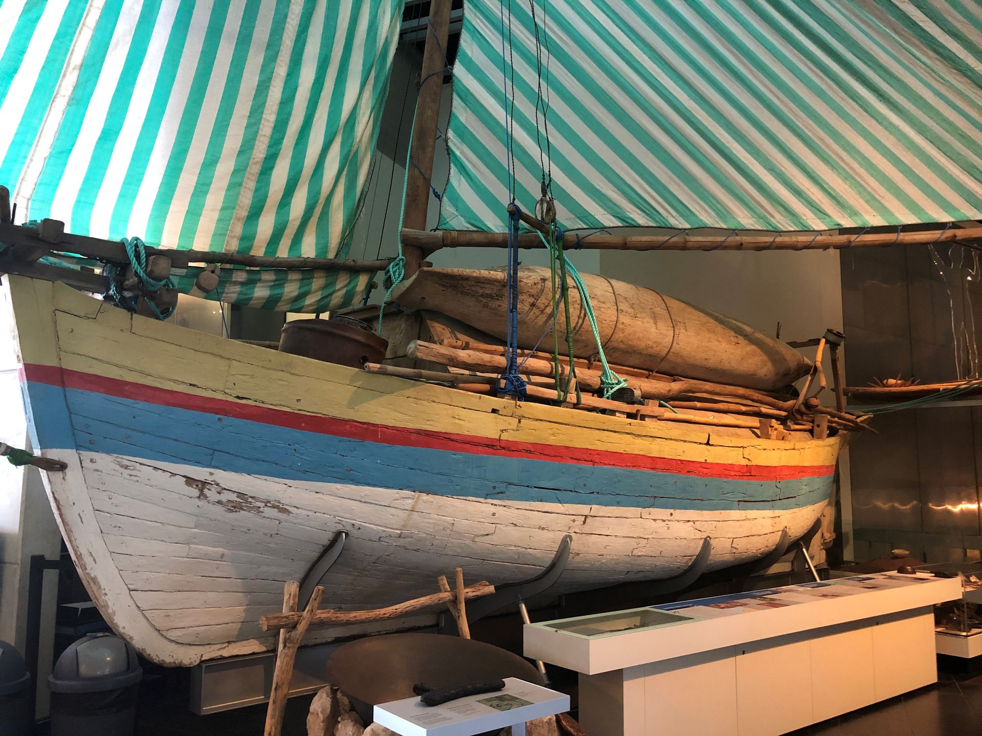 A small wooden sailing boat. The hull is painted white with yellow, red and blue stripes. The sails are striped blue and white.