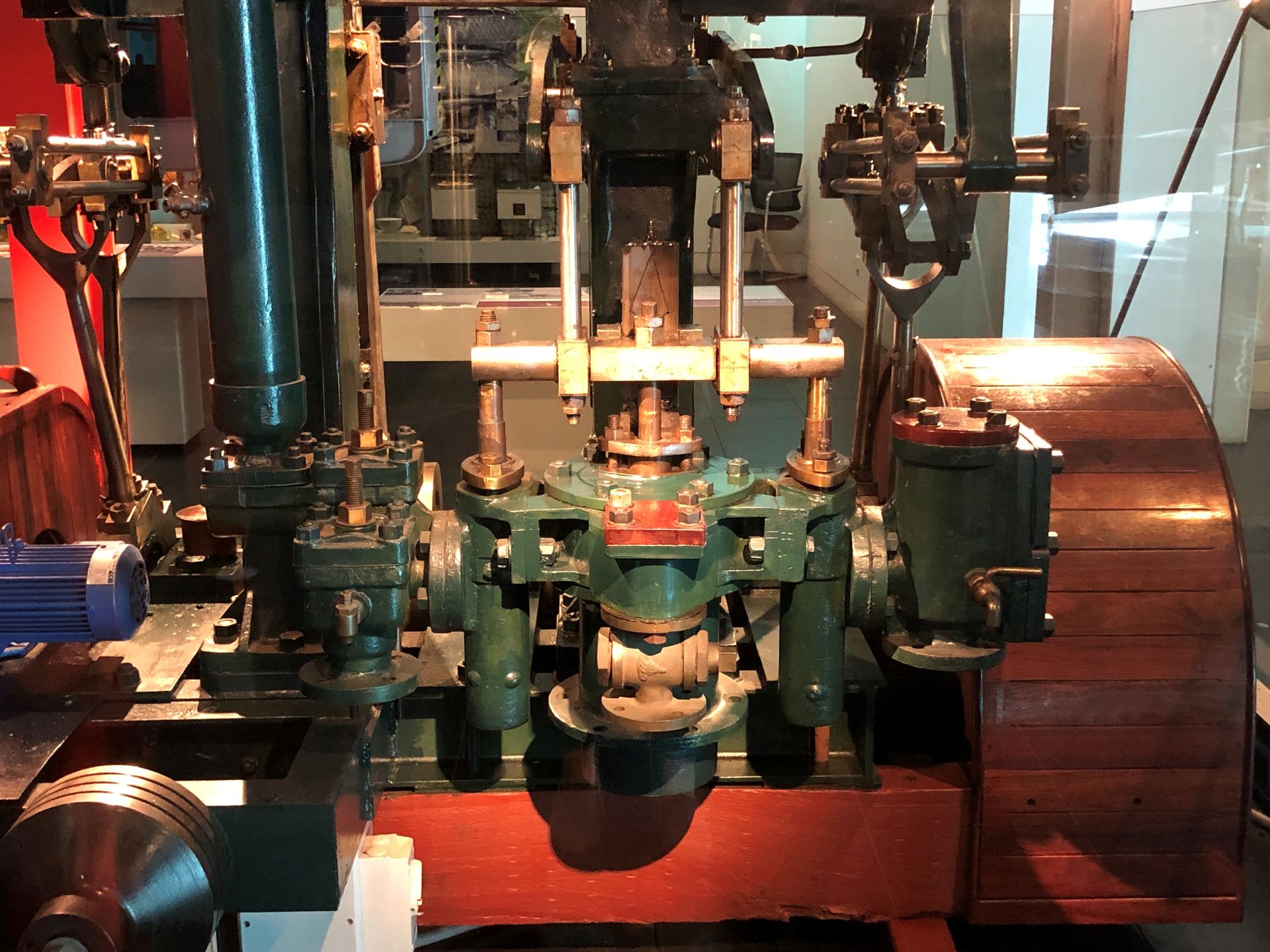 A large green engine mounted on the floor, a wooden wheel mounted vertically on its side.