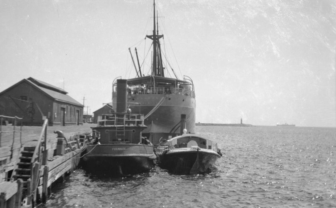 The Kangaroo towers above the Lady Forrest and the tugboat. Passengers on the Kangaroo look at the camera.