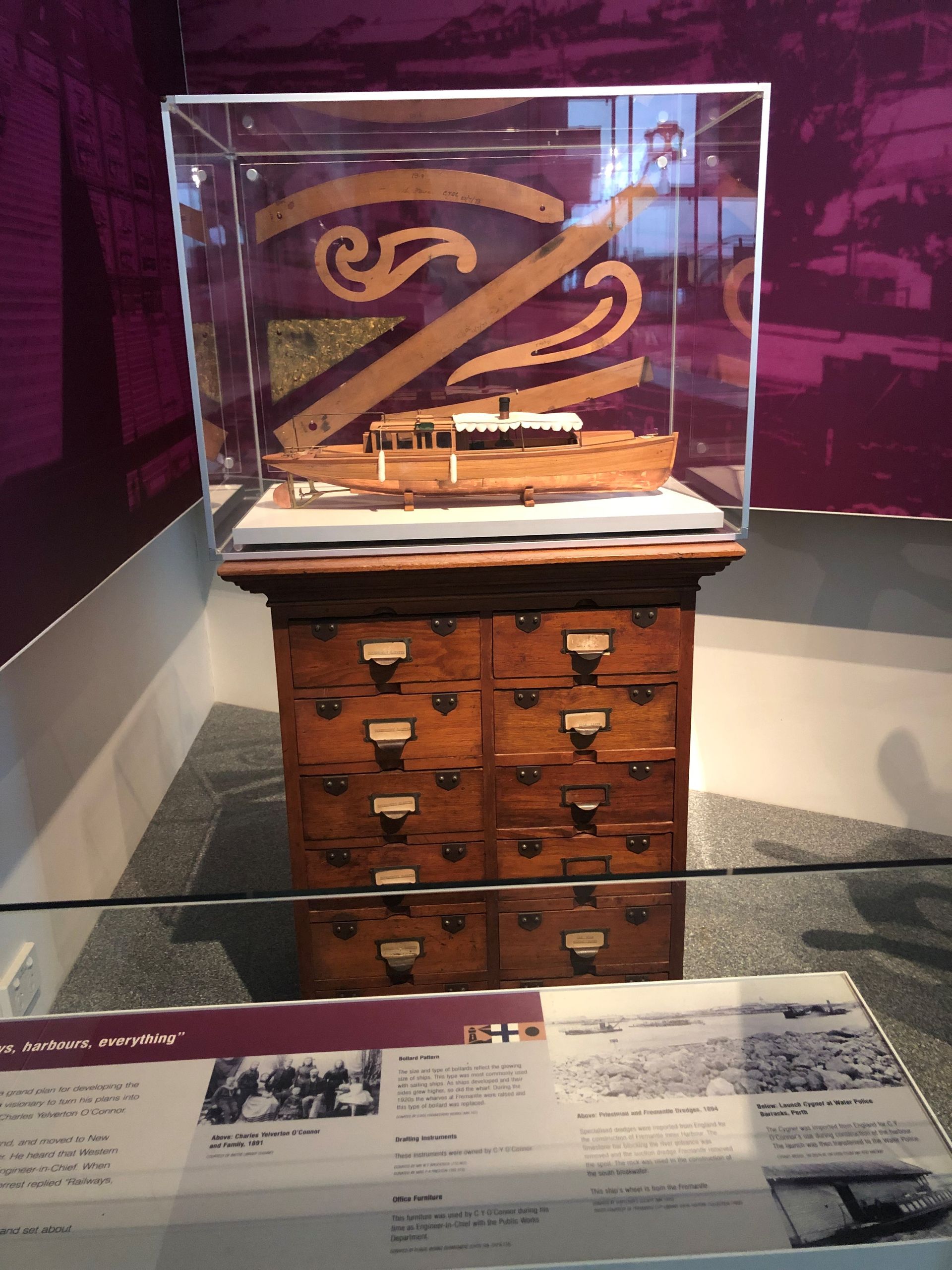 Behind a glass shield is a narrow dark wood cabinet with two columns of small drawers. On top is a glass display containing a wooden boat model.