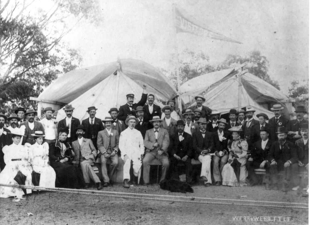 Black and white photo of a large group of white people posed in two lines, outside in front of tents.