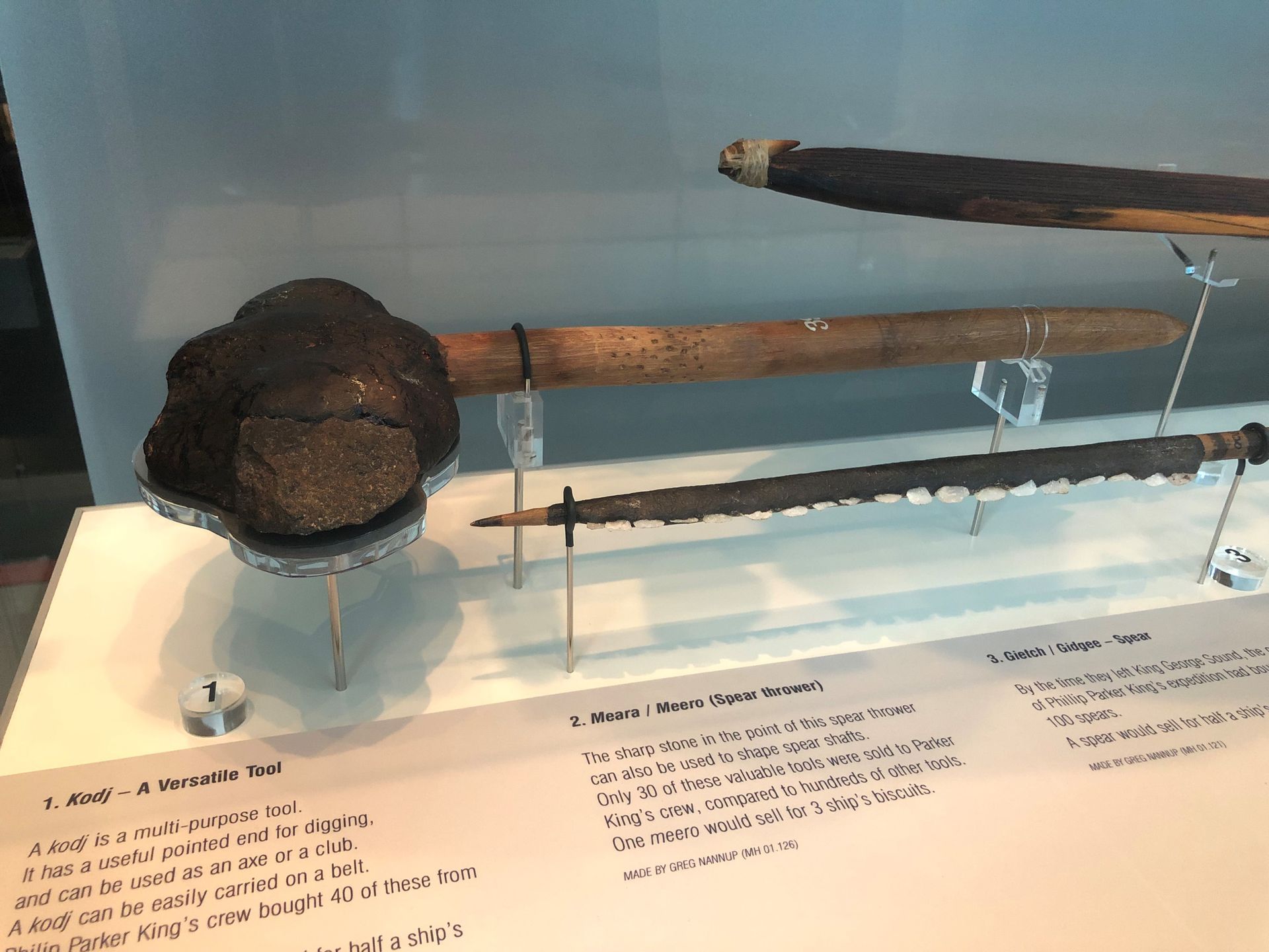 A tool with a thick wooden shaft and a very large stone head mounted horizontally behind a display case, next to a spear thrower and spear.