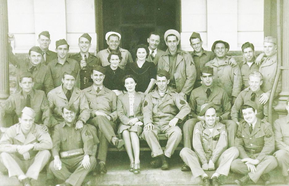 A large group of US Navy men in uniform surround three nicely dressed women, everyone is smiling at the camera.