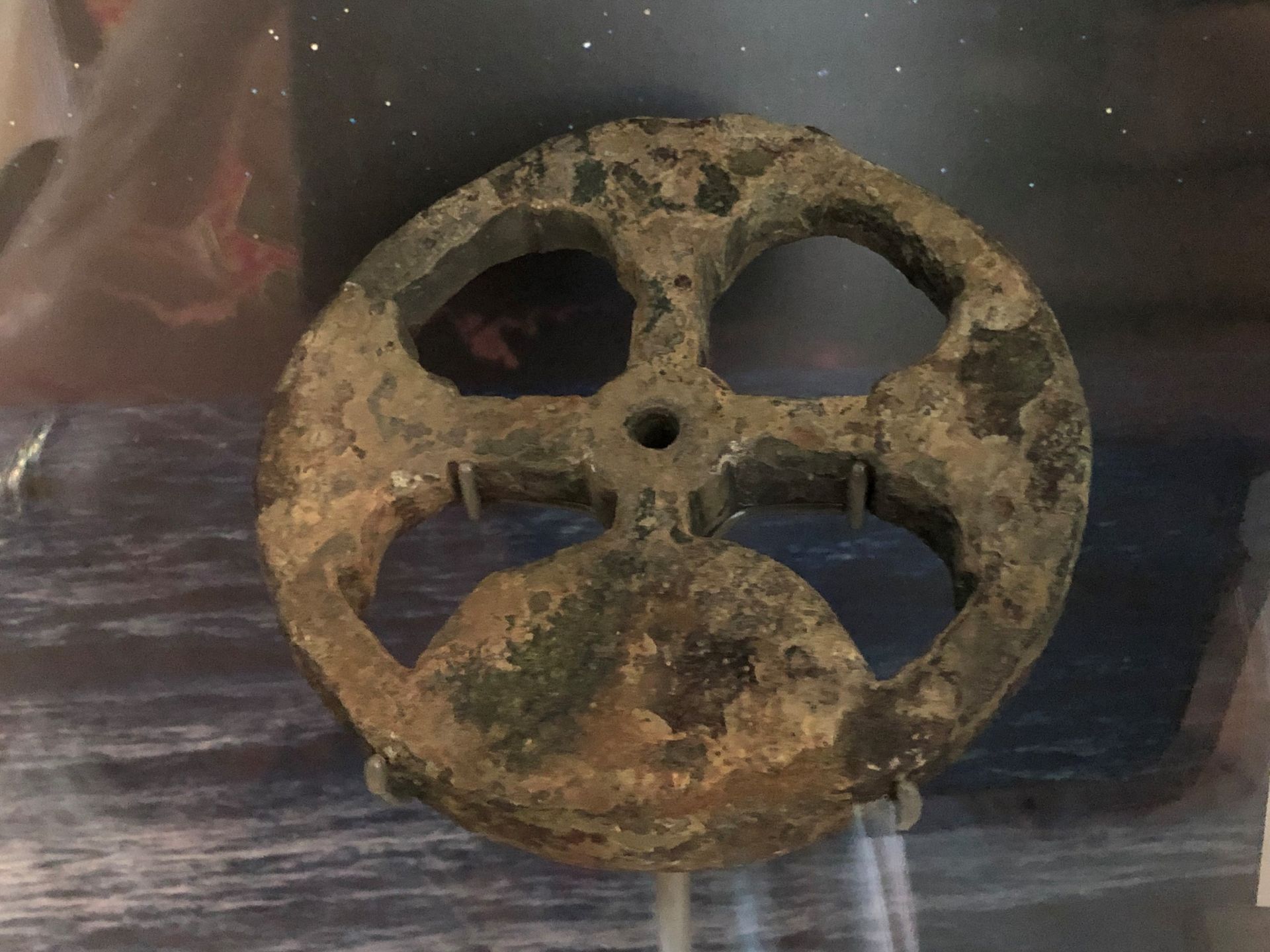 A heavily corroded metal circular object on display behind glass. There are four holes around the centre so the circle appears to be in four quarters.